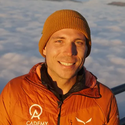 Chris Fromm im Portrait, Wollmütze und Daunenjacke, hinter ihm ein Wolkenmeer im Abendlicht.