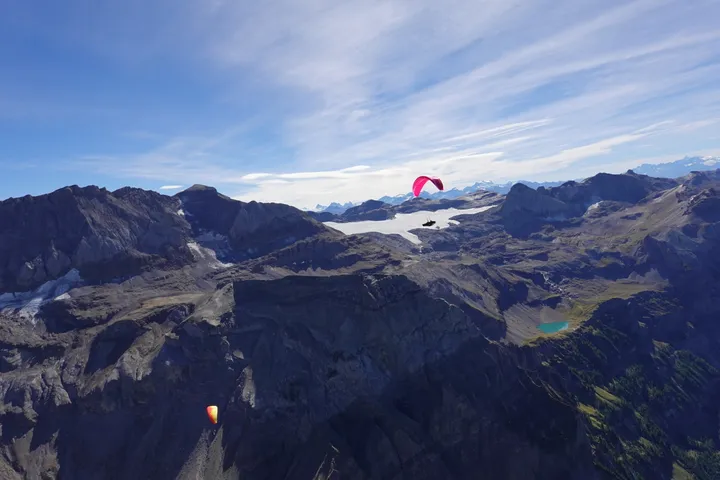 Ein pinker Gleitschirm über einer Hochebene mit zwei Bergseen, dahinter ein Gletscher und eine lange Kette verschneiter Gipfel.