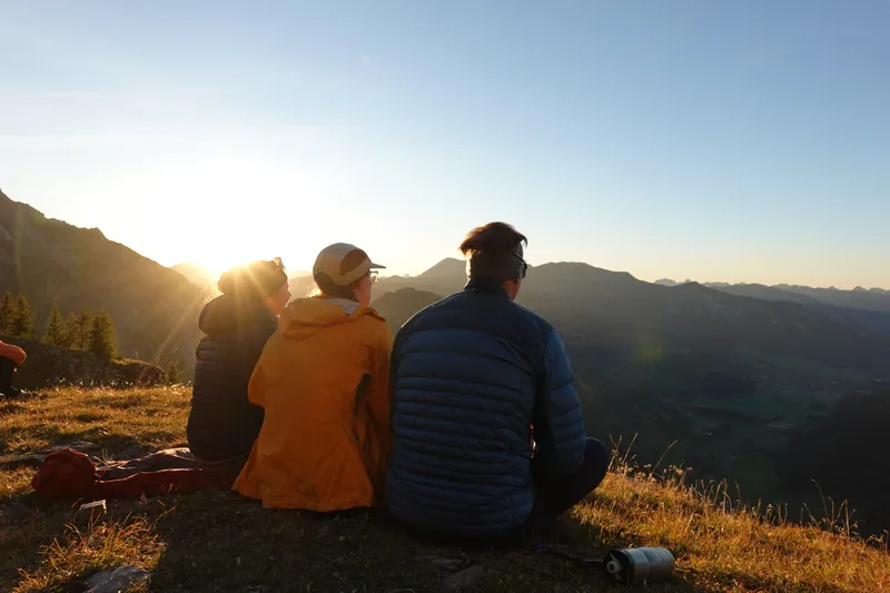 Drei Menschen sitzen am Abend auf einer Bergwiese und schauen in die untergehende Sonne.