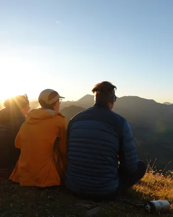 Drei Menschen sitzen am Abend auf einer Bergwiese und schauen in die untergehende Sonne.