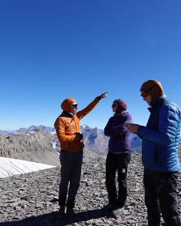 Auf einem Geröllgipfel zeigt ein Pilot in oranger Jacke in die Ferne, zwei andere schauen mit, dahinter Gletscher und Berge.