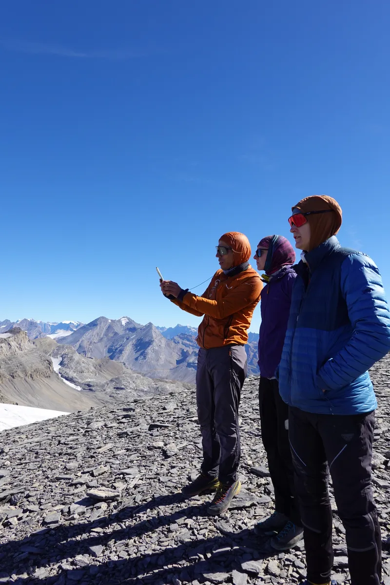 Drei Piloten auf einem Geröllgipfel, einer zeigt mit ausgestrecktem Arm über die Gletscher in die Ferne.