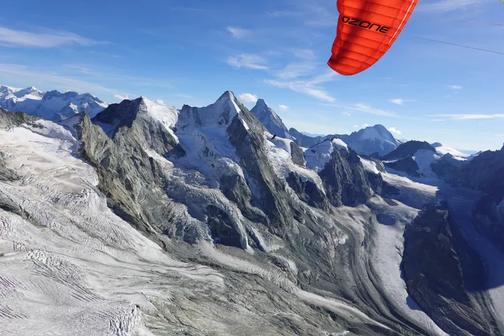 Die Spitze eines roten Gleitschirms oben im Bild, darunter vergletscherte Gipfel und Grate im Hochgebirge.