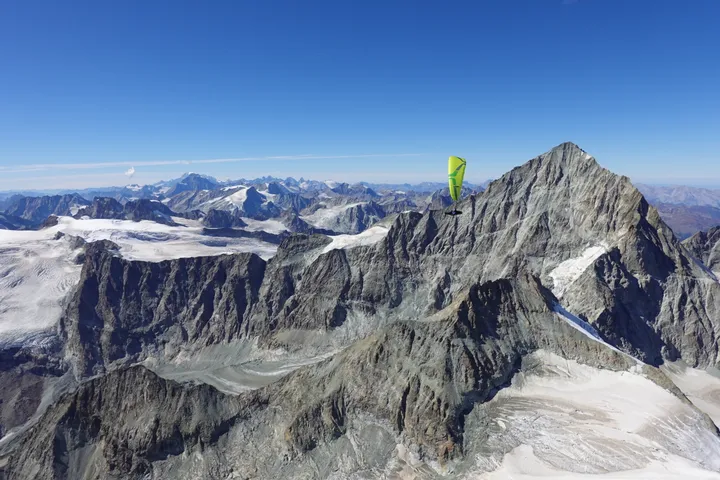 Ein gelbgrüner Gleitschirm über einem felsigen Gipfel, darunter Gletscher und Grate, am Horizont die Walliser Alpen.