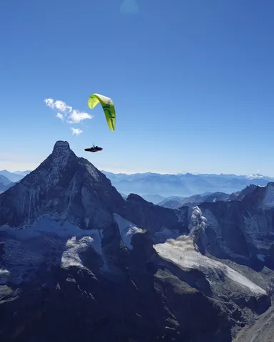 Ein gelbgrüner Gleitschirm hoch über dem Gipfel des Matterhorns, ringsum Gletscher und Viertausender unter blauem Himmel.