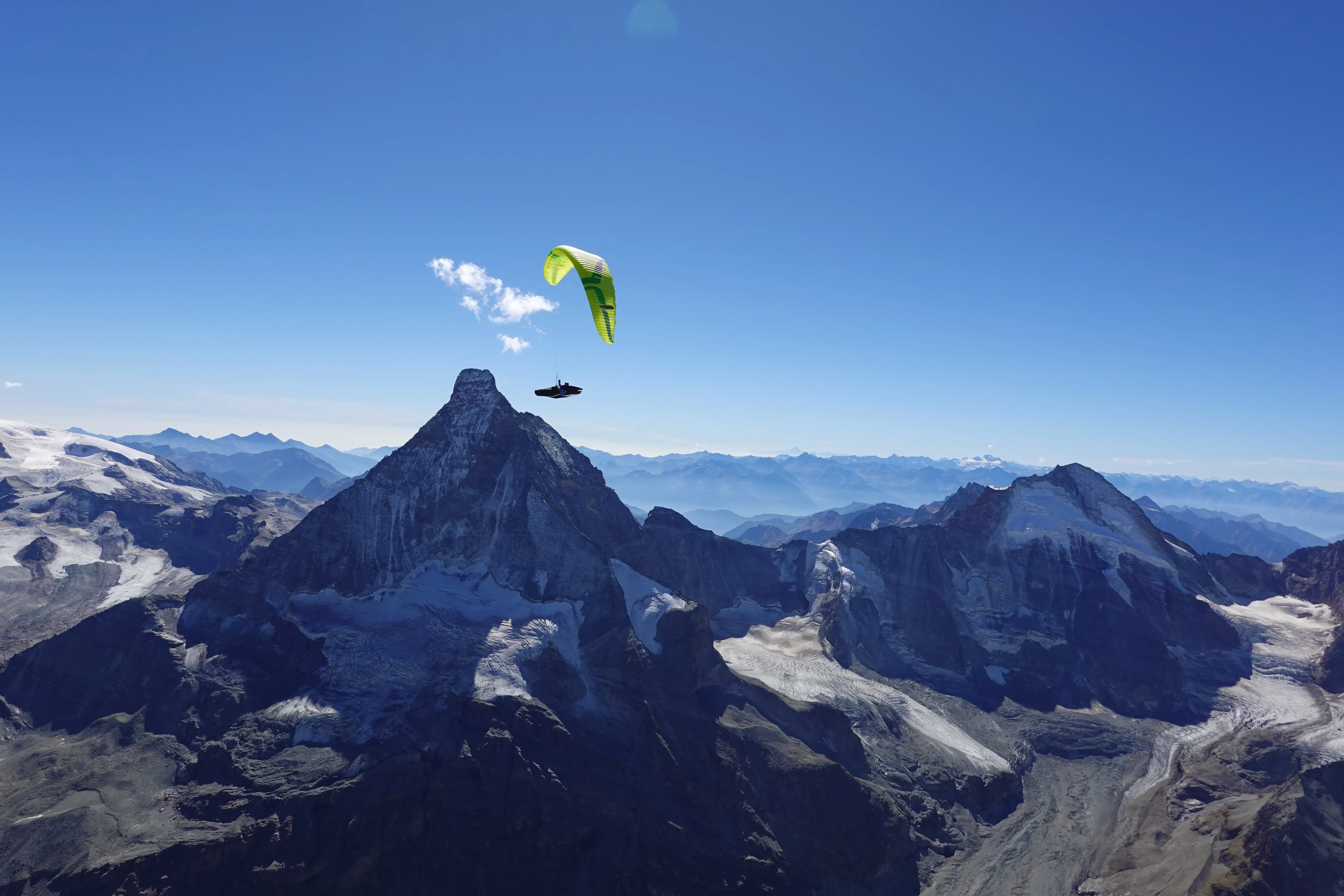 Ein gelbgrüner Gleitschirm hoch über dem Gipfel des Matterhorns, ringsum Gletscher und Viertausender unter blauem Himmel.
