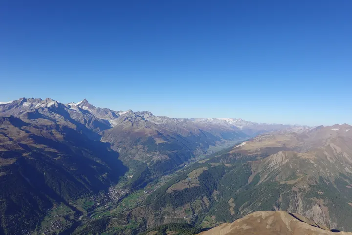 Blick aus der Luft ins Goms, ein langes grünes Tal zwischen hohen Bergen, vorne ein brauner Grat.