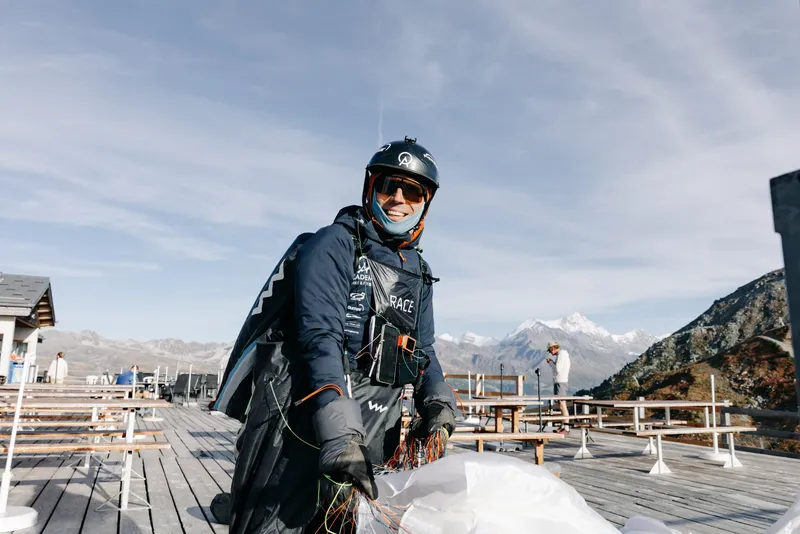 Chris Fromm im Flugoverall auf der Terrasse einer Bergstation, die Leinen des gepackten Schirms in den Händen, dahinter verschneite Gipfel.