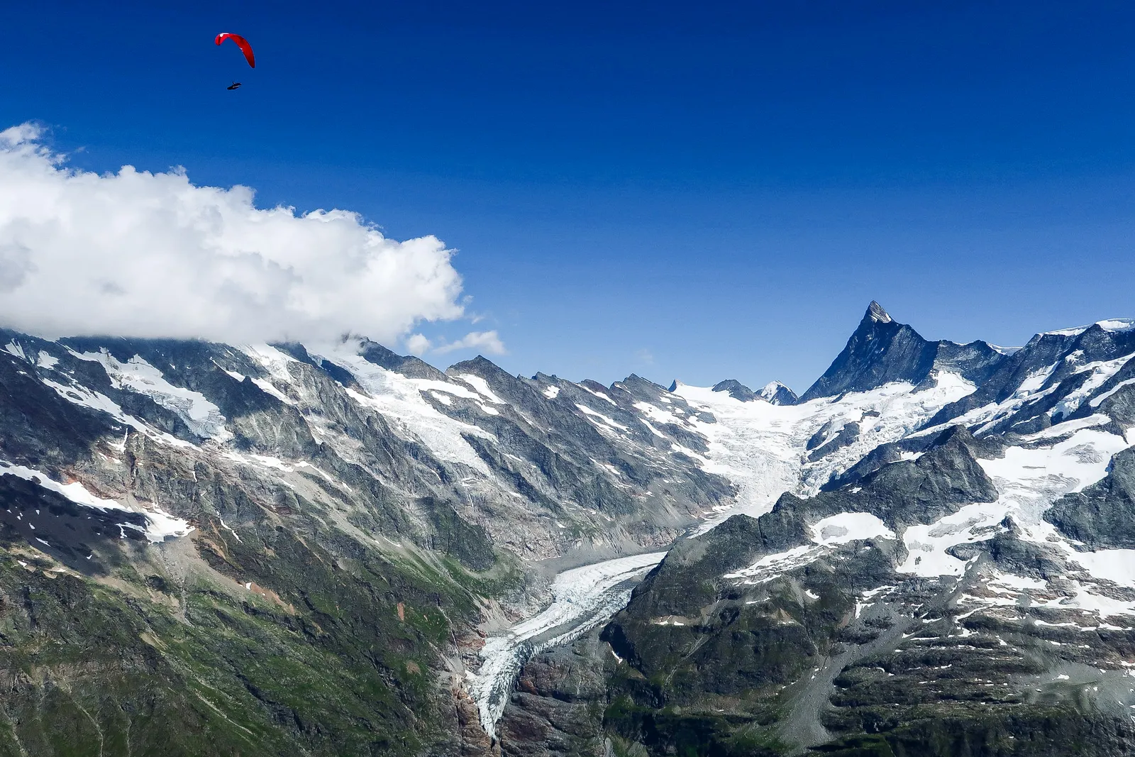 Ein roter Gleitschirm hoch über einem Gletscherbecken, dahinter verschneite Viertausender.