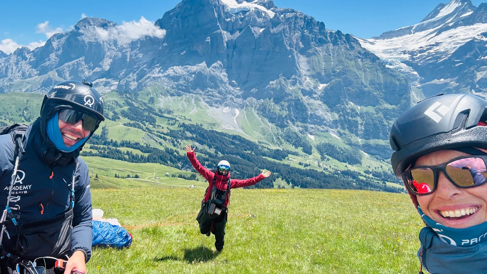 Drei Pilotinnen und Piloten auf der Startwiese, lachend, dahinter eine grosse Felswand mit Gletscher.