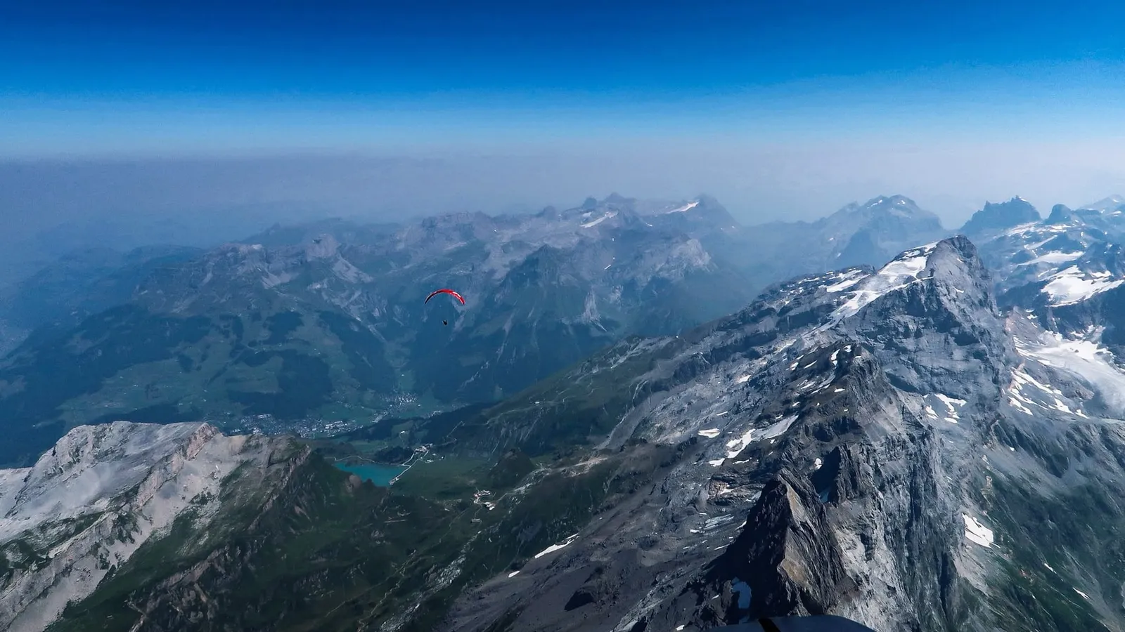 Weiter Blick über ein Tal: ein einzelner roter Gleitschirm auf dem Weg zur nächsten Bergkette, rechts ein schroffer Grat, im Dunst dahinter die weiteren Ketten.